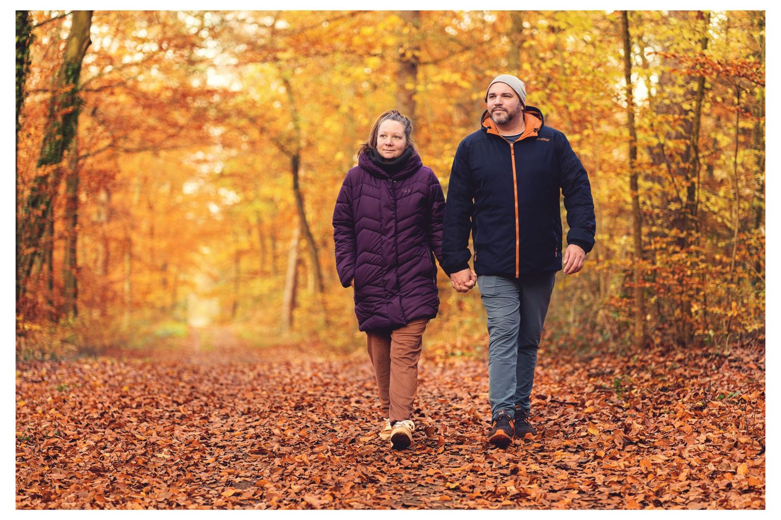A man and woman in padded jackets walk hand in hand along a leaf-covered path through autumn woodland