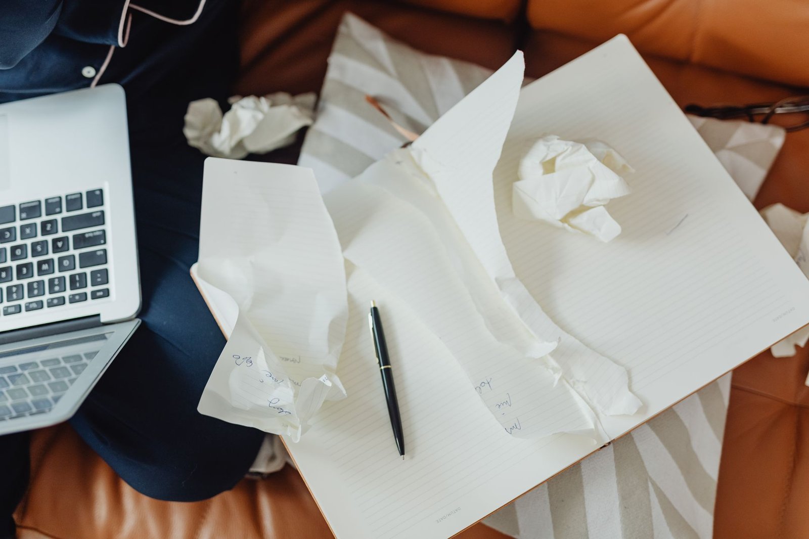Open notebook with a pen and crumpled paper on a tan sofa, next to a laptop and a person's legs in dark trousers