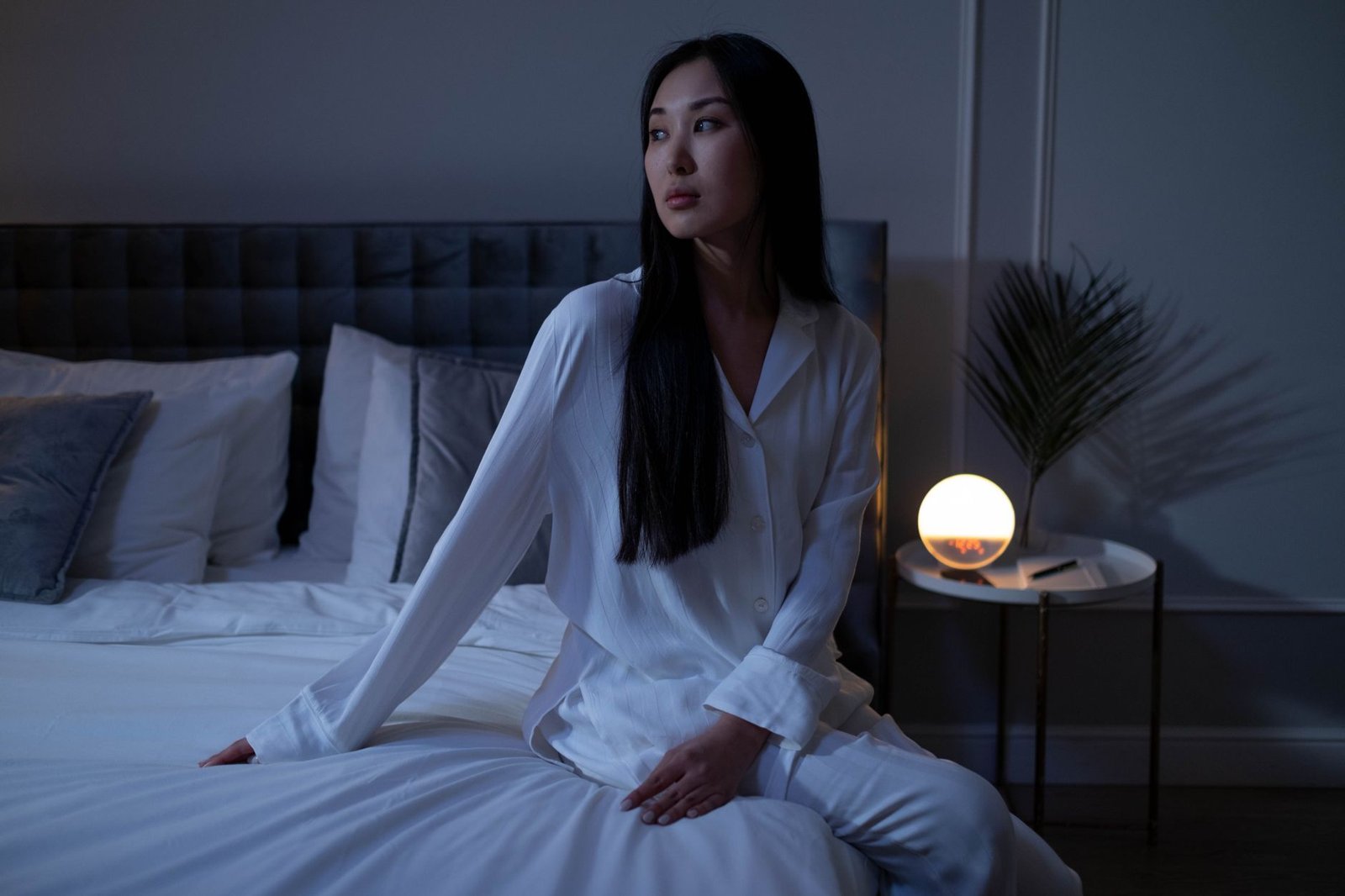 A woman in white pyjamas sits on the edge of a bed in a dim bedroom, looking away beside a softly glowing lamp